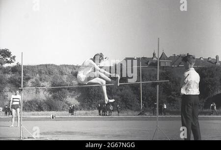 1960s, historique, athlétisme, sports scolaires inter-comté, un écolier faisant le saut en pieds-de-pieds, en utilisant la technique traditionnelle des ciseaux, regardé par un autre garçon, en gardant son œil sur le bar, Devon, Angleterre, Royaume-Uni. Lorsque les ciseaux sautent, l'athlète passe au-dessus de la barre en position assise en ramenant la jambe arrière vers le haut tandis que la jambe principale descend de l'autre côté de la barre. Bien que le saut de ciseaux ait été remplacé par de nouvelles techniques, il est resté un style utilisé par de nombreuses femmes cavaliers, avec le record du monde des femmes dans les 1960s toujours tenu par une variante des ciseaux. Banque D'Images