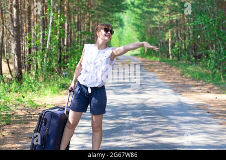 Belle jeune femme touriste avec une valise sur une route forestière lors d'une journée ensoleillée d'été sur fond d'arbres verts. Gros plan. Mise au point sélective Banque D'Images