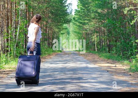 Belle jeune femme touriste avec une valise sur une route forestière lors d'une journée ensoleillée d'été sur fond d'arbres verts. Gros plan. Mise au point sélective Banque D'Images