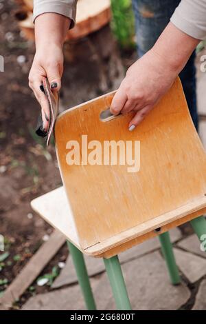 les mains des femmes en gros plan décollez la vieille peinture d'une petite chaise haute avec du papier de verre sur le fond d'un jardin d'été Banque D'Images