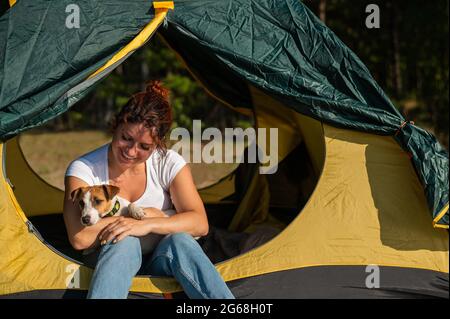 Belle femme aux cheveux rouges se repose dans la nature avec son animal de compagnie. Une fille s'assoit dans une tente touristique et encadre un Jack Russell Terrier dans la forêt. Le chien et Banque D'Images
