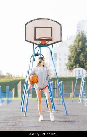 Brunette jeune femme gaie vêtue de vêtements modernes et décontractés posant avec le basket-ball au stade. Banque D'Images