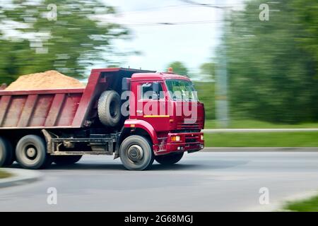 Camion-citerne industriel lourd rouge avec sable à l'arrière, panoramique flou tourné en mouvement en ville flou sur feuillage vert flou d'arrière-plan avec Banque D'Images