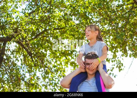 Frère défoqué de la sœur de circonscription à l'arrière. Portrait d'une fille heureuse sur les épaules de l'homme, porcgyback. Petite mouche. Famille jouant à l'extérieur. Arrière-plan de l'arbre vert. Banque D'Images