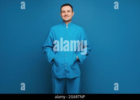 Un médecin en uniforme médical sourit et regarde la caméra sur fond bleu. Jeune médecin chirurgien isolé sur fond bleu Banque D'Images