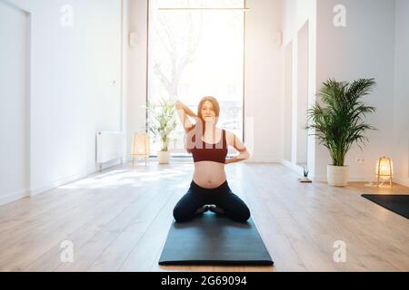 Image ensoleillée d'une femme enceinte assise sur un tapis à genoux, dos droit, pratiquant le yoga seul dans un grand studio. Il lui a pris les doigts Banque D'Images