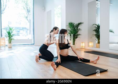 Instructeur aidant la femme enceinte à pratiquer le yoga, redressant son dos. La femme est assise sur un tapis avec des jambes étalées dans un grand studio. Elle porte Banque D'Images