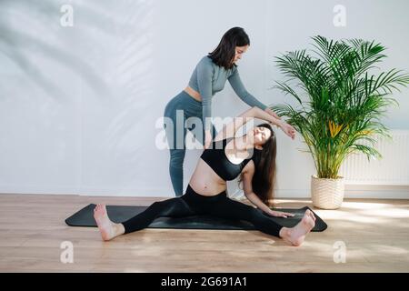 Entraîneur aidant la femme enceinte à pratiquer le yoga, ils font des étirements latéraux profonds. La femme est assise sur un tapis avec des jambes étalées dans un grand studio. Elle est wea Banque D'Images