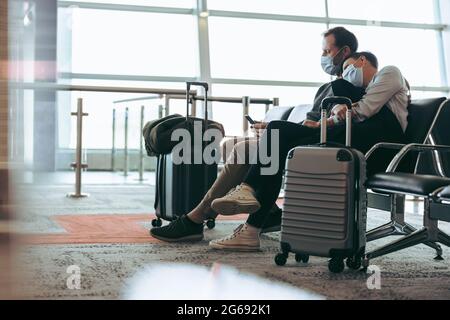 Couple portant des masques de visage assis au terminal de l'aéroport et attendant le vol. Homme utilisant un téléphone portable assis à côté d'une femme à l'aéroport pendant la corona Banque D'Images