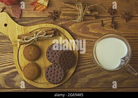 Biscuits aux pépites de chocolat sur un plateau et lait dans une tasse. Cuisson avec des bâtonnets de cannelle sur fond en bois. Tasse en forme de coeur. Composition d'automne. Banque D'Images