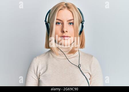 Jeune femme blonde qui écoute de la musique à l'aide d'écouteurs avec une expression sérieuse sur le visage. Simple et naturel regarder la caméra. Banque D'Images