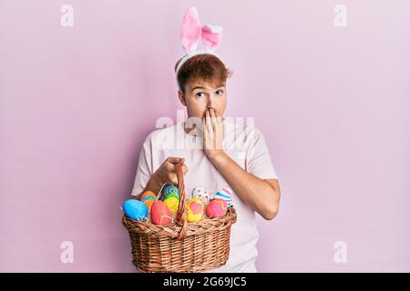 Jeune homme caucasien portant des oreilles de lapin de pâques mignonnes portant un panier en osier avec des œufs de couleur couvrant la bouche avec la main, choqué et peur de l'erreur. S Banque D'Images