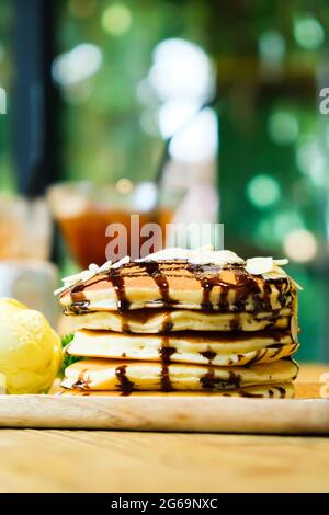 Crêpes à la banane sucrée avec garniture aux noix de macadamia et au sirop de chocolat sur plat de crêpe, posez sur une assiette en bois, jardin vert flou en arrière-plan Banque D'Images