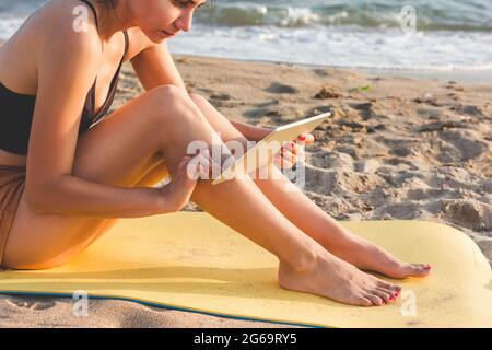Jeune femme assise sur un tapis de yoga jaune, utilisant une tablette sur la plage. Concept de communication sans fil et connexion Internet dans les endroits les plus éloignés Banque D'Images