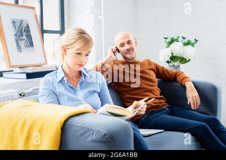 Belle femme lisant un livre tandis que son mari assis à côté de lui sur le canapé et parlant avec quelqu'un sur son téléphone mobile. Banque D'Images