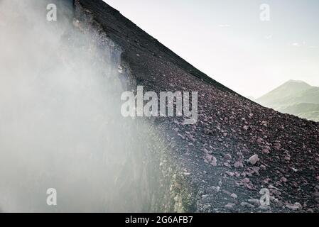 Vue rapprochée du bord d'un volcan actif, avec de la fumée de soufre qui s'élève de l'intérieur du cratère. En arrière-plan, un ciel clair et des volcans verts. Banque D'Images