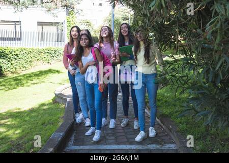 Groupe de jeunes filles Latina marchant le long d'un chemin dans un parc ou une cour d'école à l'arrière de l'école avec des cahiers et des sacs à dos. Éducation et tee Banque D'Images