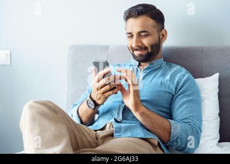 Jeune homme indien souriant utilisant un téléphone portable assis sur le lit à la maison Banque D'Images