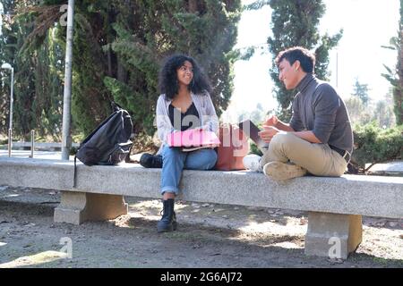 Deux étudiants latins souriant étudiant ensemble assis sur un banc à l'extérieur. La vie universitaire sur le campus. Banque D'Images