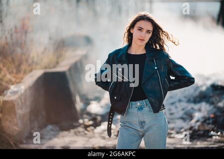 Portrait extérieur d'une belle jeune fille brune dans la veste en cuir noir avec de la fumée en arrière-plan Banque D'Images