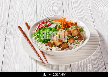 bol à poke de tofu avec riz basmati, haricots édamames, radis, concombre finement coupé et carottes dans un bol blanc avec baguettes, vue horizontale de l'abov Banque D'Images