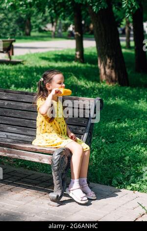 Une fille pleine longueur vêque d'une robe jaune est assise sur le bord d'un banc dans un parc d'été et mange du maïs jaune. Festival de la récolte et du maïs Banque D'Images