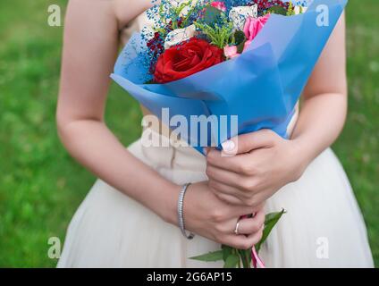 Les mains de la mariée dans la robe de mariage tiennent un bouquet de fleurs colorées de roses dans le papier d'emballage bleu, gros plan. Banque D'Images