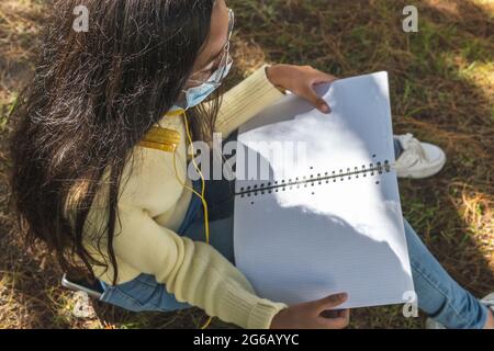 Latina adolescente portant des lunettes jaunes et des écouteurs avec masque facial tient un carnet assis dans l'herbe pendant les cours de plein air. Coronavirus pré Banque D'Images