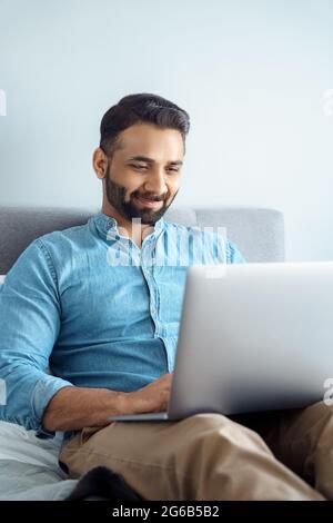 Homme d'affaires indien souriant ayant une conversation vidéo informelle sur ordinateur portable, asseyez-vous sur le lit Banque D'Images