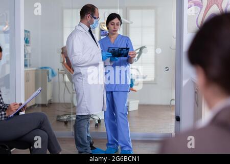 Dentiste dans la salle d'attente en stomatologie tenant une radiographie du patient expliquant le diagnostic à l'infirmière dans un couloir encombré avec la forme de remplissage du patient. Personnel de Dentistiry à la réception. Banque D'Images