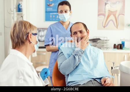 Homme malade ayant des symptômes de douleur dentaire tenant la main sur la joue tout en parlant avec un médecin senior. Patient expliquant le problème dentaire à la stomatologiste femme assise dans le bureau de clinique de l'hôpital de stomatologie Banque D'Images