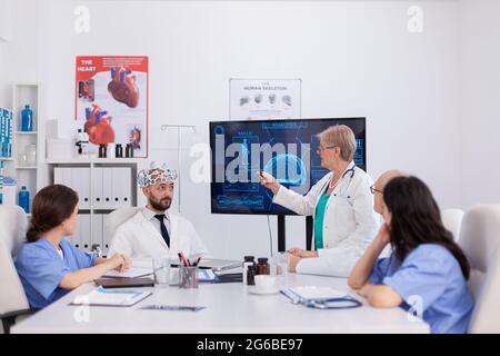 Neurologue spécialiste, femme senior, pointant vers la radiographie cérébrale, discutant de l'expertise en matière de maladie à l'aide d'un micro-casque avec capteurs dans la salle de réunion. Travail d'équipe en milieu hospitalier analyse de la présentation de brainst Banque D'Images