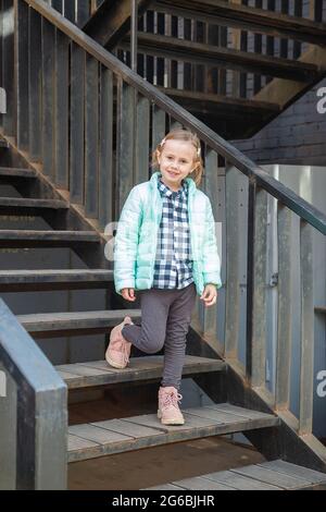 Petite fille blonde caucasienne dans un jeune tendance chaud vêtements de dessus de printemps se tient sur l'escalier de fer vintage regardant la caméra. Très beau Banque D'Images