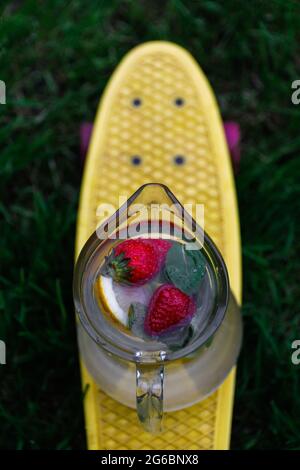 Flou artistique en verre de la limonade à la fraise, trancher le citron et les feuilles de menthe sur le panneau jaune de penny, planche à roulettes. Arrière-plan d'herbe flou. Pichet de Banque D'Images