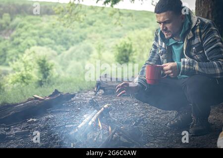 Un homme avec une tasse de boisson chaude se réchauffe par le feu dans la forêt. Banque D'Images