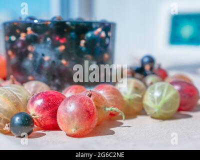 Baies de cassis dans un verre d'eau avec le cube de glace. Les groseilles à maquereau colorées sont dispersées autour de la tasse. Fenêtre et coque en arrière-plan. Banque D'Images