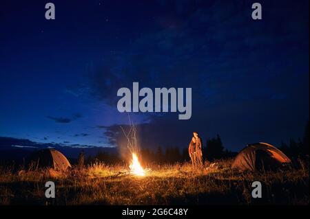 Magnifique paysage de nuit ciel nuageux sur une colline herbeuse avec tentes de camp, feu de camp et randonneurs. Les jeunes voyageurs, hommes et femmes, profitent de la vue sur le ciel bleu de nuit tout en se tenant près des tentes touristiques. Banque D'Images