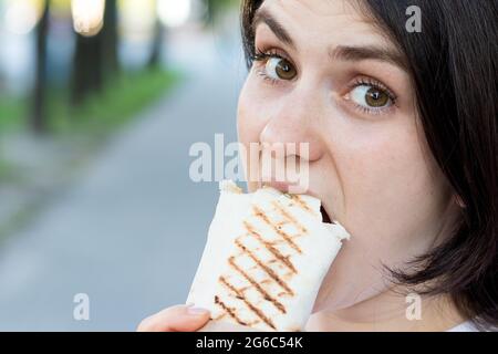 La femme Brunette surmange le shawarma dans une rue de la ville. Rouleau de pita de restauration rapide de rue avec de la viande et des légumes. Placer pour le texte. Banque D'Images