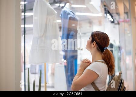 Femme caucasienne regardant une fenêtre de magasin et rêvant d'une robe. Banque D'Images