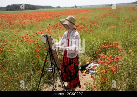 Femme mature artiste peint une image sur un champ de fleurs des coquelicots Portrait de l'artiste au travail.vue latérale. Banque D'Images