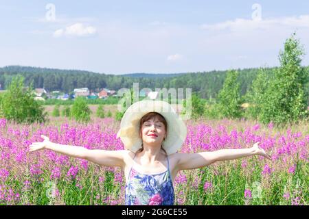 Jeune femme dans un champ de fleurs dans un chapeau de paille sur un fond de forêt verte et ciel bleu lors d'une chaude journée d'été. Mise au point sélective Banque D'Images