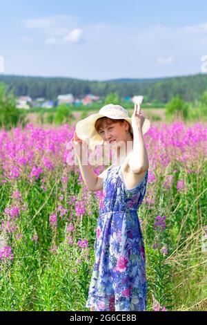 Jeune femme dans un champ de fleurs dans un chapeau de paille sur un fond de forêt verte et ciel bleu lors d'une chaude journée d'été. Mise au point sélective Banque D'Images