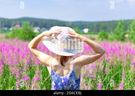 Jeune femme dans un champ de fleurs dans un chapeau de paille sur un fond de forêt verte et ciel bleu lors d'une chaude journée d'été. Mise au point sélective Banque D'Images