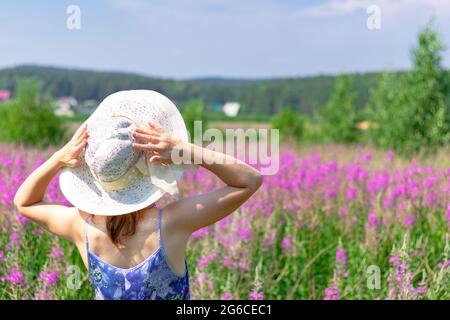 Jeune femme dans un champ de fleurs dans un chapeau de paille sur un fond de forêt verte et ciel bleu lors d'une chaude journée d'été. Mise au point sélective Banque D'Images