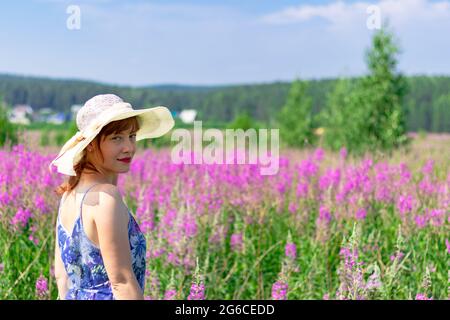 Jeune femme dans un champ de fleurs dans un chapeau de paille sur un fond de forêt verte et ciel bleu lors d'une chaude journée d'été. Mise au point sélective Banque D'Images