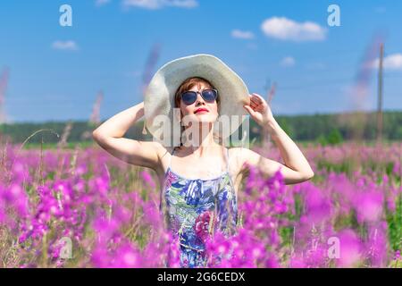 Jeune femme dans un champ de fleurs dans un chapeau de paille sur un fond de forêt verte et ciel bleu lors d'une chaude journée d'été. Mise au point sélective Banque D'Images