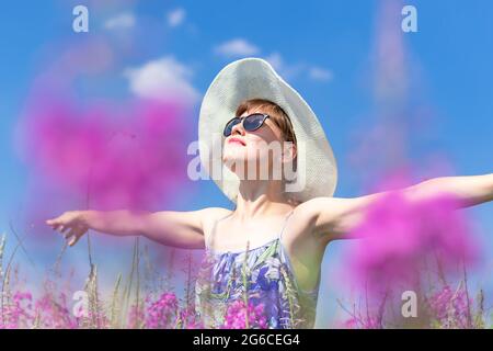 Belle jeune femme dans un chapeau et des lunettes de soleil dans un champ de fleurs lors d'une chaude journée d'été contre un ciel bleu. Mise au point sélective. Gros plan Banque D'Images