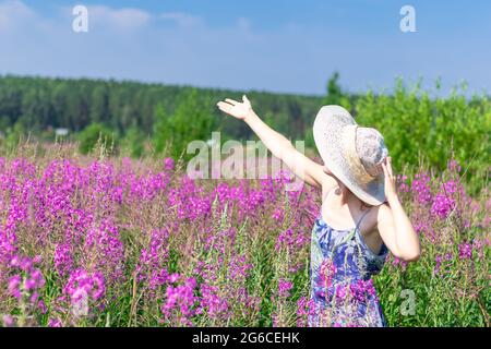 Jeune femme dans un champ de fleurs dans un chapeau de paille sur un fond de forêt verte et ciel bleu lors d'une chaude journée d'été. Mise au point sélective Banque D'Images