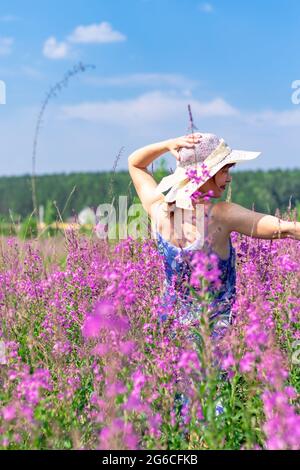 Jeune femme dans un champ de fleurs dans un chapeau de paille sur un fond de forêt verte et ciel bleu lors d'une chaude journée d'été. Mise au point sélective Banque D'Images