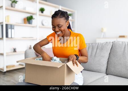 Service de livraison en ligne. Une femme afro-américaine joyeuse qui déballe sa boîte à colis à la maison Banque D'Images